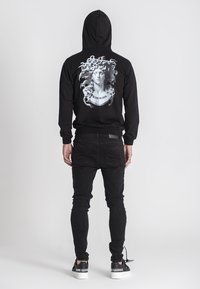 Crni dukserica s grafičkim printom koji prikazuje poprsje i lepršavu kosu na leđima. U kombinaciji s crnim uskim farmerkama i kontrastnim tenisicama.