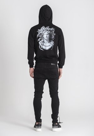 Crni dukserica s grafičkim printom koji prikazuje poprsje i lepršavu kosu na leđima. U kombinaciji s crnim uskim farmerkama i kontrastnim tenisicama.