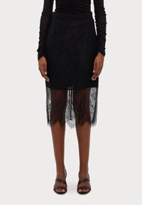 Black lace skirt featuring a fitted upper layer and sheer lace hem with scalloped edges. Worn with black strappy high heels.