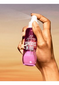 Przezroczysta różowa butelka mgiełki perfum z białą dyszą spray, z napisem "SUNDAYS IN RIO" w białym kolorze.