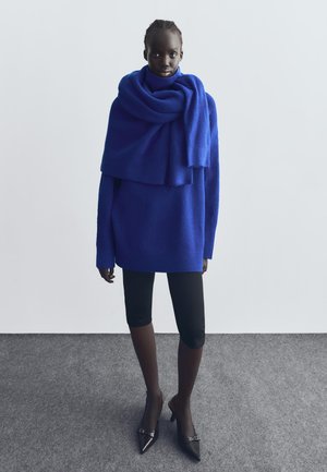 Kék kötött pulóver magas nyakkal és lezser anyaggal, fekete térd alatti leggingsszel és hegyes fekete magassarkú cipővel párosítva.