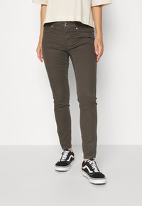 Jeans skinny-fit de cor verde oliva, com cinco bolsos, fecho de botão e uma textura suave. Combinados com sapatilhas pretas e brancas.