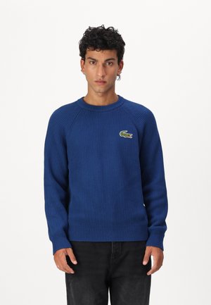 Blauw geribbeld gebreid sweater met een ronde hals en raglanmouwen. Bevat een Lacoste krokodillenlogo op de linkerborst. Draagt samen met donkere jeans.