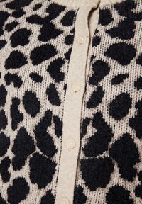 Cardigan crème avec imprimé léopard noir, présentant un tricot texturé et une fermeture à boutons sur le devant. Tissu doux avec détails de motif visibles.