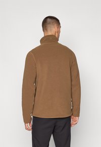Brun fleece genser med høy krage, teksturert stoff og kontraststing. Har en liten logo ved halsen for detalj.