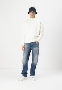 Sweatshirt crème, jean bleu déchiré et baskets blanches. Le modèle porte un chapeau seau noir. Design simple avec une coupe décontractée.