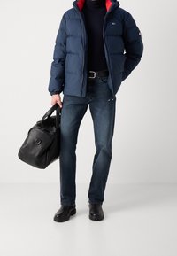 Marinblå puffjacka med röd foder, mörka jeans, svart läderväska och svarta stövlar. Jackan har dragkedja fram och elastiska ärmslut.