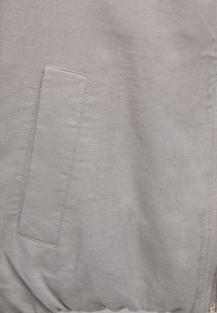 Gros plan sur un tissu gris clair avec une poche rectangulaire et des détails de couture visibles sur un vêtement.