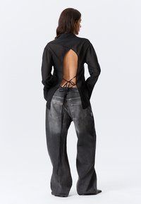 Chemise noire transparente à manches longues avec une grande découpe dans le dos, nouée à la taille. Associée à un jean gris délavé, ample, avec des poches arrière.