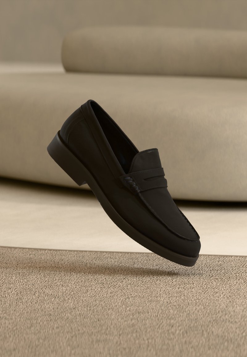 Mocassin en cuir noir flottant au-dessus d'un tapis beige avec un canapé beige en tissu en arrière-plan.