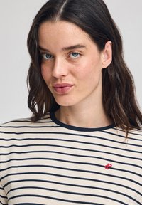 Randig t-shirt i marinblått och krämfärgat, rund halsringning och liten röd logotyp på bröstet, tillverkad av mjukt bomullstyg.