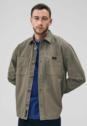 Camicia color khaki con bottoni, due tasche frontali, minuteria in argento e colletto classico, indossata sopra una t-shirt blu. Tessuto in cotone liscio.