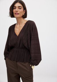 Blusa plissettata marrone scuro con scollatura a V, cintura elastica e maniche ampie. Indossata con pantaloni marroni a vita alta.