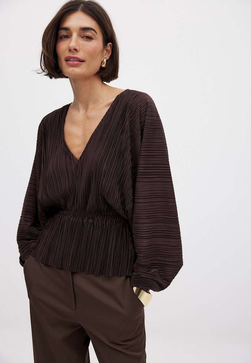 Blusa plissettata marrone scuro con scollatura a V, cintura elastica e maniche ampie. Indossata con pantaloni marroni a vita alta.