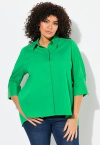 Camisa de color verde brillante, de manga larga y con botones, con cuello puntiagudo, corte holgado y aberturas laterales; combinada con jeans de mezclilla oscura.