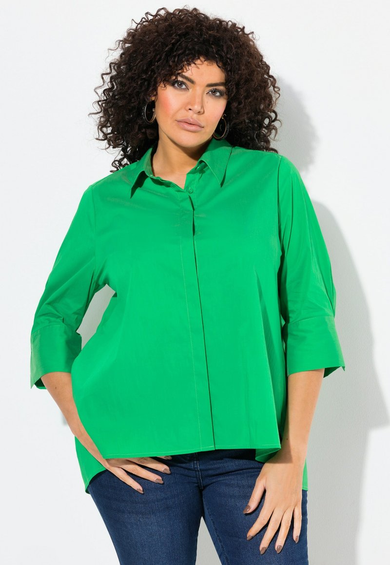 Camisa de color verde brillante, de manga larga y con botones, con cuello puntiagudo, corte holgado y aberturas laterales; combinada con jeans de mezclilla oscura.