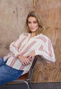Femme aux cheveux blonds portant un chemisier rayé crème et rose et un jean bleu, s'appuyant en arrière sur une chaise marron contre un mur en bois.