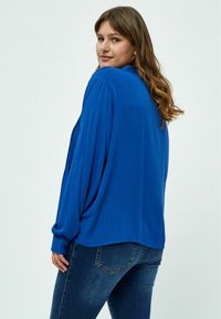 Blusa blu a maniche lunghe con design drappeggiato e schiena plissettata, abbinata a jeans di denim scuro. Presenta polsini con bottone e una vestibilità comoda.