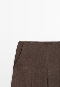 Gros plan sur la ceinture d'un pantalon en tissu marron texturé avec poches latérales et une coupe avant plate sur fond blanc.