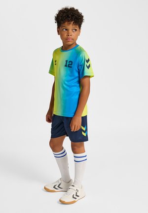 Jongen met krullend haar, gekleed in een blauw-gele sporttrui en marineblauwe korte broek, staand en zijwaarts kijkend tegen een witte achtergrond.
