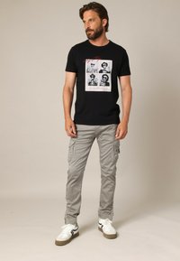 Homme avec une barbe portant un t-shirt graphique noir, un pantalon cargo gris clair et des baskets blanches, se tenant devant un fond beige.