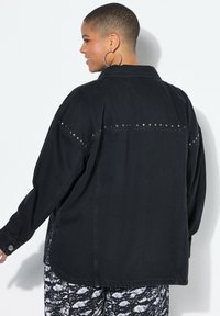 Giacca di jeans nera oversize con borchie argentate lungo la parte posteriore, dotata di colletto a camicia e maniche lunghe. Abbinata a pantaloni a fantasia.