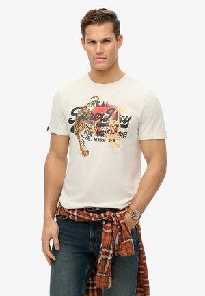White cotton t-shirt featuring a tiger graphic, black lettering, and red circular accent. Paired with denim jeans and a tied plaid shirt.