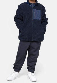 Enfant portant une veste en polaire bleu marine avec une poche poitrine zippée, un pantalon de jogging bleu marine et des baskets blanches, debout les mains dans les poches.