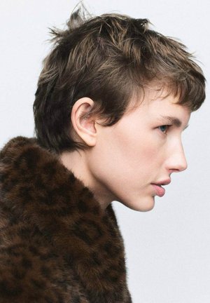 Capa corta de piel marrón con cuello de estampado de leopardo, que presenta una textura de pelo natural y un tono de piel suave, mostrada de perfil contra un fondo claro.