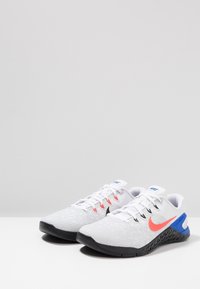 Zapatillas deportivas blancas con parte superior texturizada, detalles en rosa y azul, suela y cordones negros. Logotipo de Nike visible en el lateral y en la lengua.