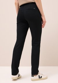 Person, der schwarze, eng anliegende Hosen mit zwei Gesäßtaschen und weiße Sneakers mit schwarzen "V"-Logos trägt, steht auf einem beigen Boden.