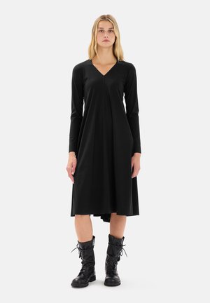 Vestito nero a maniche lunghe con scollatura a V, forma svasata e lunghezza fino al ginocchio. Indossato con stivali neri con lacci, creando un contrasto di stile.