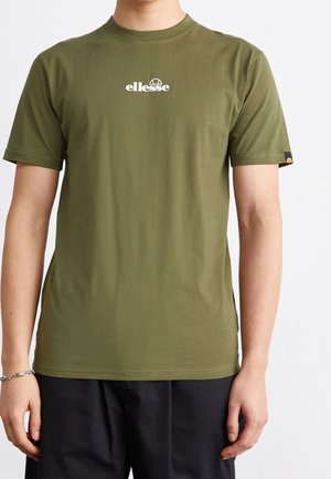 Personne portant un t-shirt olive vert avec le logo ellesse sur la poitrine et un pantalon noir, se tenant contre un fond blanc uni.