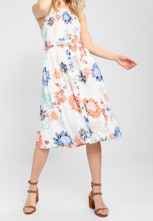 Femme portant une robe midi blanche sans manches avec des motifs floraux orange, bleu et violet, et des sandales marron à talons blocs ornées de clous.