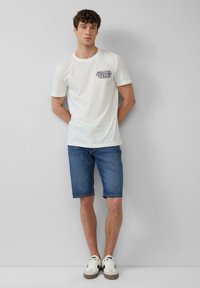T-shirt en coton blanc avec un texte graphique bleu sur la poitrine gauche, associé à un short en denim bleu et des baskets blanches. Texture lisse, design décontracté.