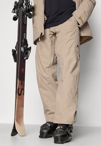 Beige skibroek gemaakt van waterafstotend materiaal, met een losse pasvorm, verstelbare tailleband en logo-detail op de dij. Skis en laarzen inbegrepen.
