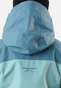 Light blue hooded jacket featuring a darker blue panel at the shoulders, SWEDEMOUNT logo embroidered in navy, smooth texture, and waterproof material.