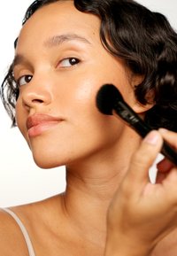 Pinceau à maquillage avec une tête ronde noire appliquant du blush sur une peau lisse et bronzée. L'arrière-plan est neutre, mettant en valeur l'application du produit.