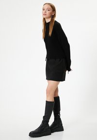 Schwarzer, gerippter Pullover mit ausgefranstem Saum, kombiniert mit einem schwarzen Minirock. Das Model trägt knielange schwarze Stiefel mit klobigen Sohlen.