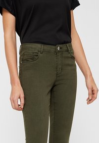 Jeans verde oliva in tessuto misto cotone, caratterizzati da una vita media, tasche anteriori e posteriori, e chiusura con zip e bottone.