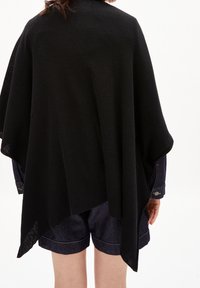 Svart stickad poncho, lättviktig, löst sittande design med en texturerad yta. Hänger över axlarna och har en asymmetrisk fåll.