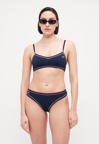 Mornarsko moder bikini set s tankimi naramnicami in kontrastnim rdečim obrobom. Ima prilegajoč zgornji del in nizko rezanje spodnjega dela, narejen iz gladkega, elastičnega materiala.