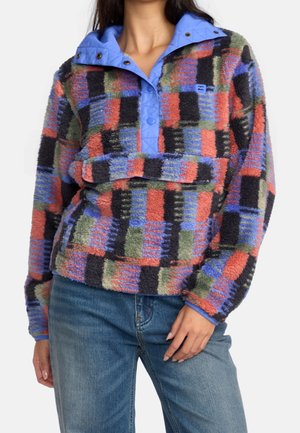 Frau trägt einen mehrfarbigen karierten Fleece-Pullover mit blauem Stehkragen und Jeans, steht mit einer Hand auf der Hüfte.