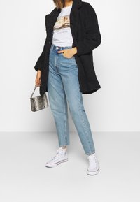 Abrigo negro y peludo sobre una camiseta gráfica; jeans de tiro alto azul claro; zapatillas blancas; bolso pequeño plateado con textura.