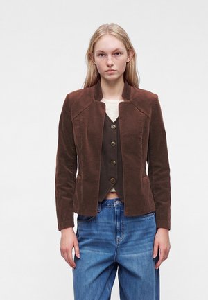 Femme portant une veste en velours côtelé marron sur un gilet marron boutonné, une chemise blanche et un jean bleu, debout devant un fond blanc uni.