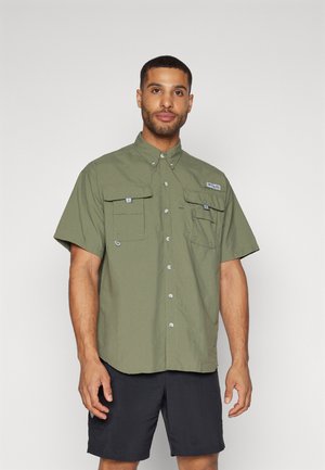 Chemise à manches courtes de couleur olive, fabriquée en tissu léger. Elle présente deux poches poitrine, une fermeture à bouton et un col.