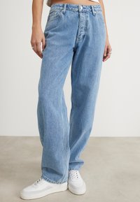 Jean en denim bleu clair avec une coupe décontractée, présentant une braguette à boutons à l'avant, des passants de ceinture et des poches latérales, associés à des baskets blanches.