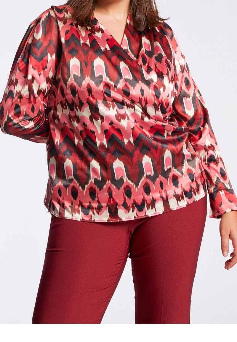 Femme portant un chemisier rouge à manches longues avec un motif et un col en V, assorti à un pantalon rouge foncé uni sur un fond uni.