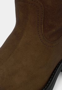 Botte de cheville en daim marron avec des coutures détaillées, un bout arrondi et une texture lisse, avec une semelle en caoutchouc contrastant avec le matériau supérieur doux.