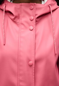 Ροζ αδιάβροχο μπουφάν με κουκούλα και κορδόνια, με τέσσερα στρογγυλά κουμπιά στον μπροστινό κεντρικό μέρος.